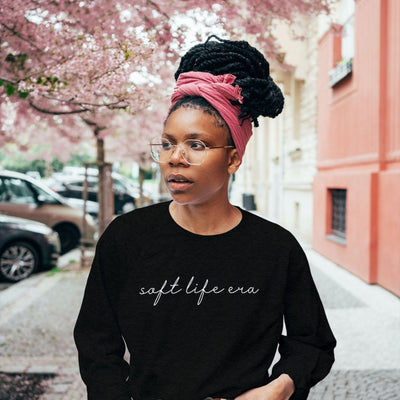 african american woman with glasses and passion twists wrapped in a bun, models the soft life cozy girl sweatshirt while walking down the street with Japanese cherry blossom tree