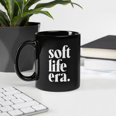 picture of soft life era glossy black mug on desk with plant, a keyboard, and a stack of books